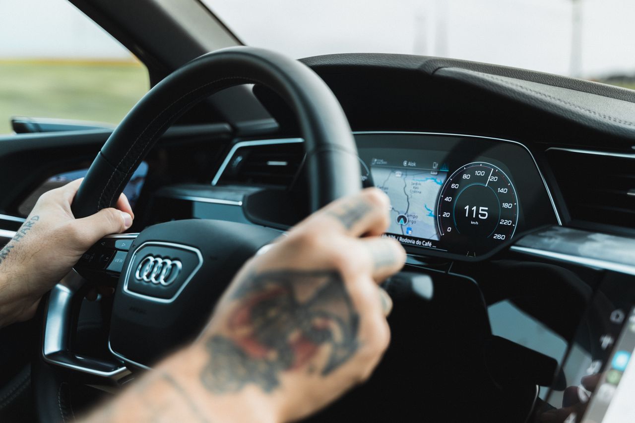 Close-up of a tattooed driver handling a car with an Audi steering wheel and digital display showing 115 km/h.